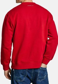 Sudadera roja con mangas largas, puños y dobladillo acanalados, que muestra "LONDON W 11" en la parte posterior del cuello. El material parece suave y texturizado.