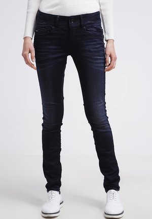 Person iført mørke skinny jeans, hvid ribbet langærmet top og hvide snøresko, stående mod en ensfarvet baggrund.