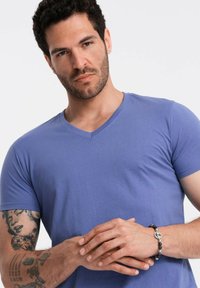 T-shirt a manica corta con scollo a V in tessuto azzurro chiaro; texture liscia. Presenta un design aderente. Accessorio: braccialetto scuro con perline e dettagli in argento.