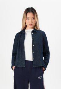 Young woman standing with hands in pockets, wearing dark denim jacket, white shirt, and navy blue pants with white and maroon stripes.