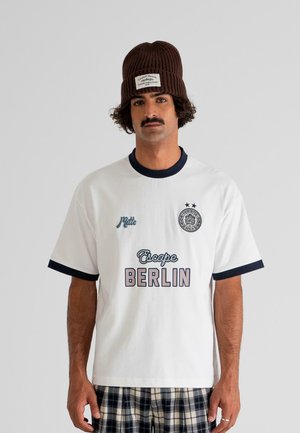 Man with mustache wearing brown knit beanie, white t-shirt with black trim and "Escape Berlin" text, and plaid shorts against plain background.