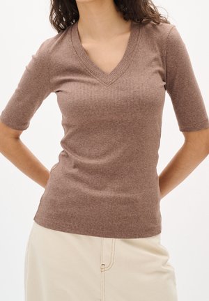Kvinde iført en brun ribbet V-hals kortærmet sweater og lyse beige bukser, stående med hænderne bag ryggen.