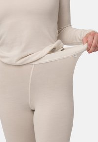 Osoba oblečená v béžových elastických legínách a odpovídajícím dlouhém tričku s dlouhým rukávem, která natahuje pas kalhot, aby ukázala pružnost látky.