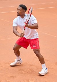 Tennisemängija kannab valget polo särki ja punaseid lühikesi pükse, hoides käes reketit savikattes. Jalatsid on valged tumesiniste detailidega.