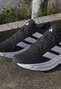 Chaussures de course Adidas noires et blanches avec des semelles blanches posées sur un pavé extérieur rugueux près d'une bordure basse en pierre.