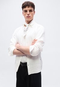 Jeune homme aux cheveux courts et bruns, portant une chemise blanche boutonnée, un foulard à motifs autour du cou, et un pantalon noir, debout les bras croisés.