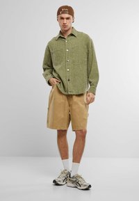 Camisa de botón verde texturizada con dos bolsillos frontales, combinada con pantalones cortos color beige. El calzado incluye zapatillas deportivas plateadas y negras.