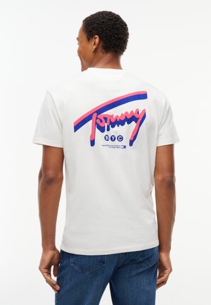 Mann trägt weißes T-Shirt mit großem pink-blauem "Tommy NYC" Logo auf dem Rücken und blauen Jeans, schaut weg von der Kamera.