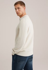 Gaišas adīts džemperis ar rakstainu tekstūru, ar polo tipa apkakli un ribotām piedurknēm, kombinēts ar tumši ziliem džinsiem.