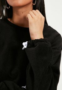 Schwarzer Fleecepullover mit gerafften Bündchen und weißem Kordelzug. Nahaufnahme des Ärmels und der Hand, die Weichheit und Textur hervorhebt.