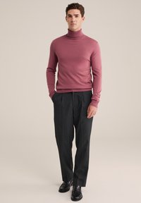 Svetlo roza turtleneck pulover iz fine pletene tkanine, kombiniran s temno sivimi nabranimi hlačami in črnimi loafers čevlji, stojijo ob nevtralnem ozadju.