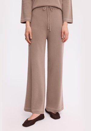 Kobieta nosząca szerokie dzianinowe spodnie taupe z paskiem ściągającym w talii oraz ciemnobrązowe plecione płaskie buty, stojąca na białym tle.