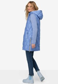 Gilet matelassé bleu clair avec une capuche, doté de poches latérales et d'une fermeture éclair, associé à un jean en denim bleu et des bottines assorties.