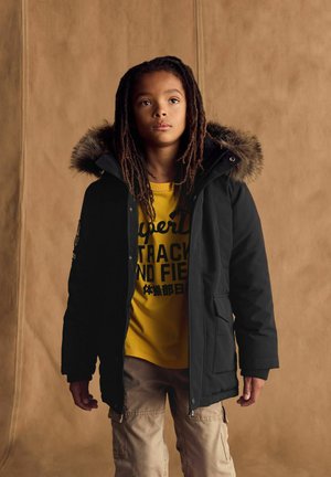 Parka noire avec capuche en fourrure, fermeture éclair et bouton, poches latérales ; porté sur un t-shirt graphique jaune, associé à un pantalon cargo beige.