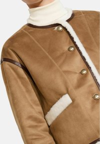 Giacca in suede marrone con colletto in pelliccia ecologica, bottoni dorati e dettagli in pelle. Presenta una tasca frontale con rifinitura in pelliccia ecologica.