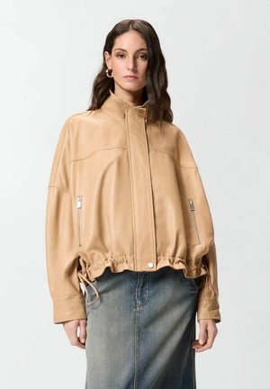 Donna che indossa una giacca in pelle color cuoio con tasche con cerniera e orlo con coulisse, abbinata a una gonna di jeans blu sbiadita, in piedi contro uno sfondo bianco.