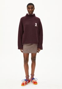 Bordeaux fleecetrui met een hoge kraag en een witte lintlogo, gecombineerd met een geweven bruine rok en kleurrijke sportschoenen.