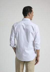 Camisa blanca de manga larga con cuello abotonado, puños enrollados y dobladillo curvado. Llevada con pantalones de lino beige.