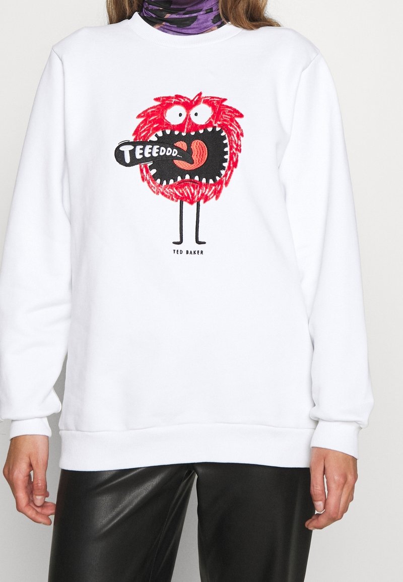 Sweat-shirt blanc avec un graphisme de monstre poilu rouge criant "TEEEDDD" dans une bulle de dialogue noire, porté avec un pantalon noir et une écharpe violette autour du cou.