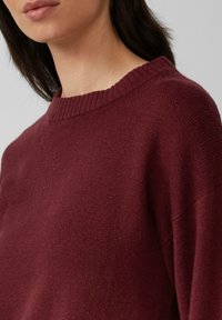 Vrouw in een geribbelde crewneck trui in diep bordeaux, waarbij de textuur en pasvorm op schouder en bovenborst zichtbaar zijn.