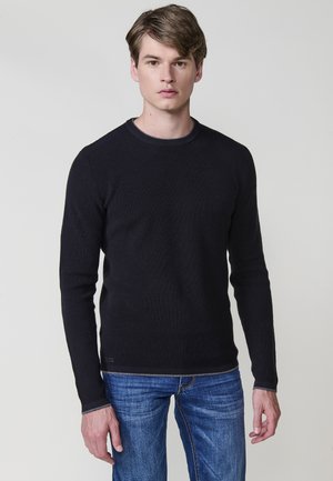 Maglione a coste nero con maniche lunghe e colletto rotondo, abbinato a jeans in denim blu. Sottotoni grigi lungo il collo e l'orlo.