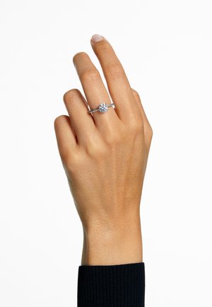 Diamanten verlovingsring met een ronde centraal gemonteerde steen, zilveren band en complexe, gestructureerde accenten langs de shank. Huidkleurige hand die het vasthoudt.