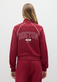 Felpa rossa con la scritta "BOSTON" in lettere bianche sulla schiena, con colletto a zip e polsini e orlo a costine. Tessuto morbido con una texture liscia.