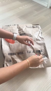 Barna kraft papírcsomag nagy narancssárga logóval és vonalkóddal, amelyet egy arany karkötőt viselő kéz nyit egy világos fa felületen.