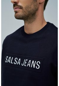 Sudadera azul marino con un parche rectangular que lleva el texto blanco "SALSA JEANS". La tela parece suave y tiene un cuello acanalado.