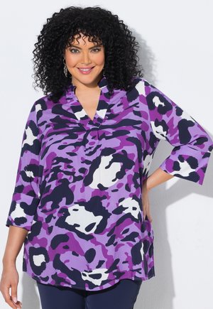 Femme souriante aux cheveux noirs bouclés porte un haut à imprimé abstrait violet, noir et blanc avec des manches 3/4, debout, une main sur la hanche.