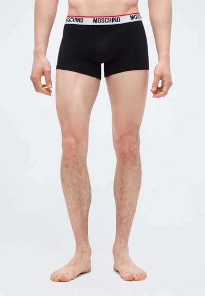Αρσενικό μοντέλο που φοράει μαύρα μποξεράκια Moschino με λευκή και κόκκινη ζώνη μέσης, όρθιο, ξυπόλυτο, σε απλό λευκό φόντο.