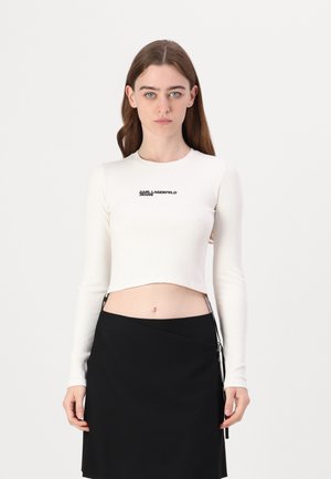 Bijela rebrasta dugorukava crop majica s logotipom "KARL LAGERFELD" u crnoj boji, u kombinaciji s crnom suknjom visokog struka s bočnim patentnim zatvaračem.