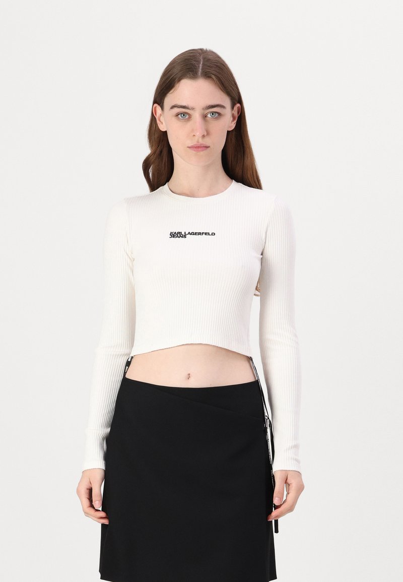 Bijela rebrasta dugorukava crop majica s logotipom "KARL LAGERFELD" u crnoj boji, u kombinaciji s crnom suknjom visokog struka s bočnim patentnim zatvaračem.