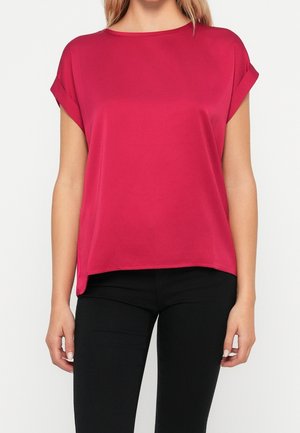 Vrouw draagt een rood, loszittend shirt met korte mouwen dat gecombineerd is met een zwarte broek, staand tegen een effen witte achtergrond.