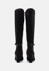 Μαύρες σουέντ μπότες μέχρι το γόνατο με διακριτική γυαλάδα, διαθέτουν φερμουάρ στο πίσω μέρος και γυαλιστερή μαύρη λωρίδα γύρω από τον αστράγαλο. Σχεδιασμός με χοντρό τακούνι.