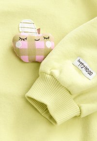 Light yellow sweater featuring a stitched apple patch in pink and green plaid. Includes ribbed cuffs and a sewn brand label.