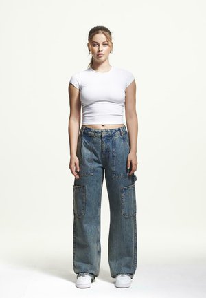 Weißes kurzärmliges Croptop, kombiniert mit übergroßen blauen Denim-Cargohosen mit großen Taschen, getragen mit weißen Sneakern vor einem schlichten Hintergrund.