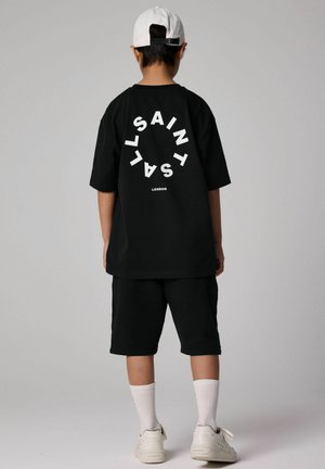 Czarny t-shirt z dużym białym napisem "SAINT" z tyłu, w połączeniu z czarnymi szortami i białymi sneakersami, z teksturowanym ribbed.