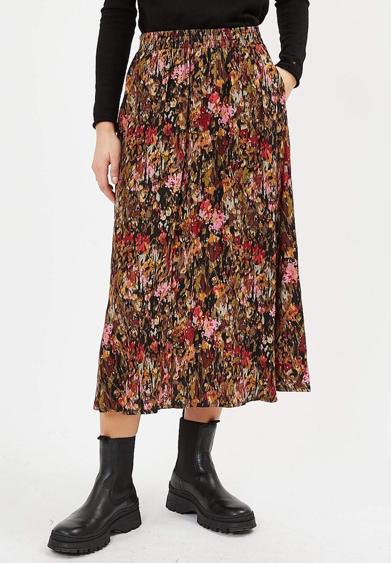 Femme portant une jupe midi plissée à motif floral multicolore, un haut noir et des bottines noires épaisses, debout les mains dans les poches.