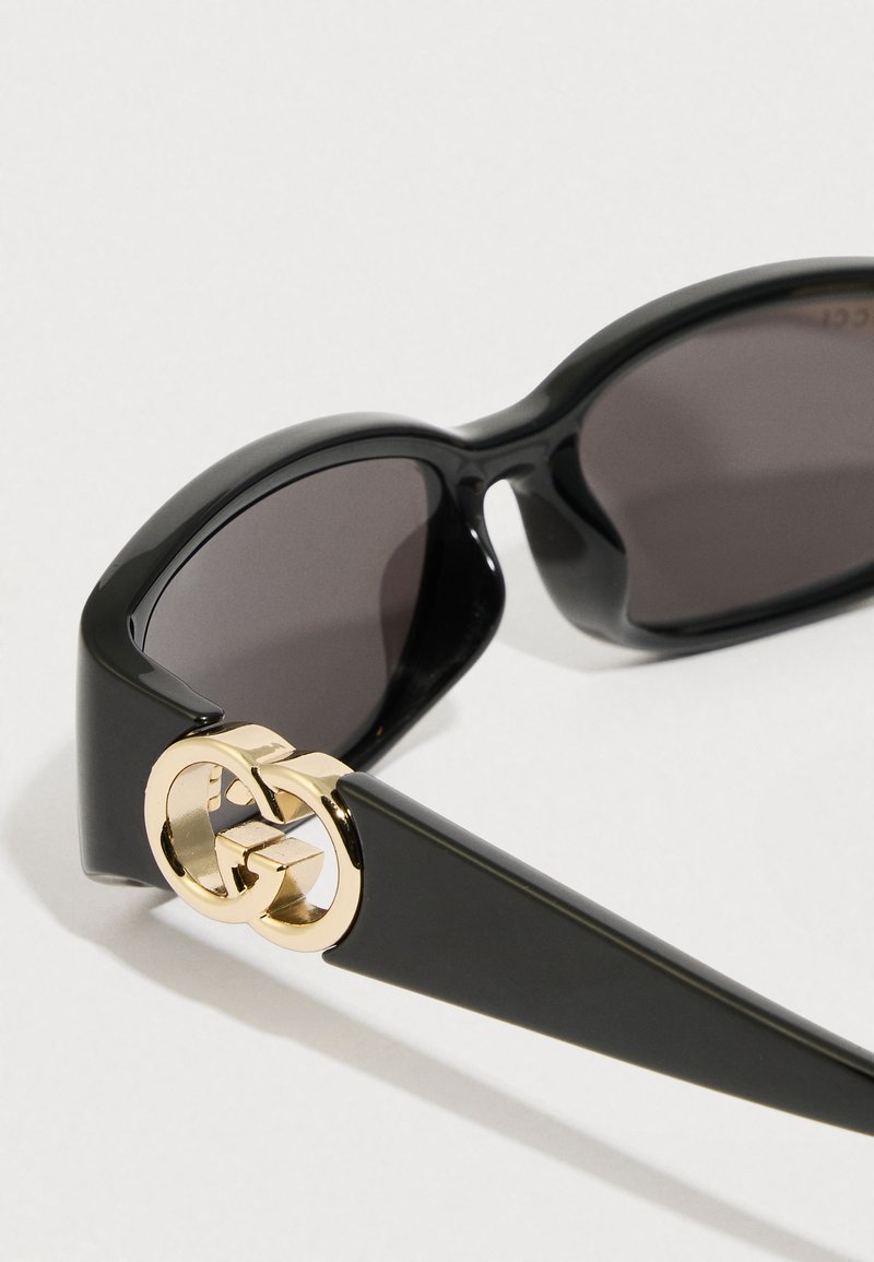 Schwarze Sonnenbrille mit glänzendem Finish, ausgestattet mit dunklen Gläsern und einem goldenen, ineinandergreifenden Logo-Akzent an den Bügeln.