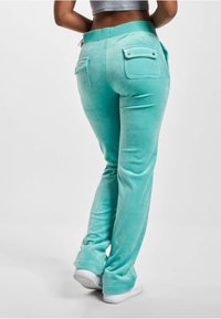 Turkusowe spodnie z weluru z dopasowanym paskiem, dwiema tylnymi kieszeniami z zapięciami na guziki i gładką fakturą, zestawione z białym obuwiem.