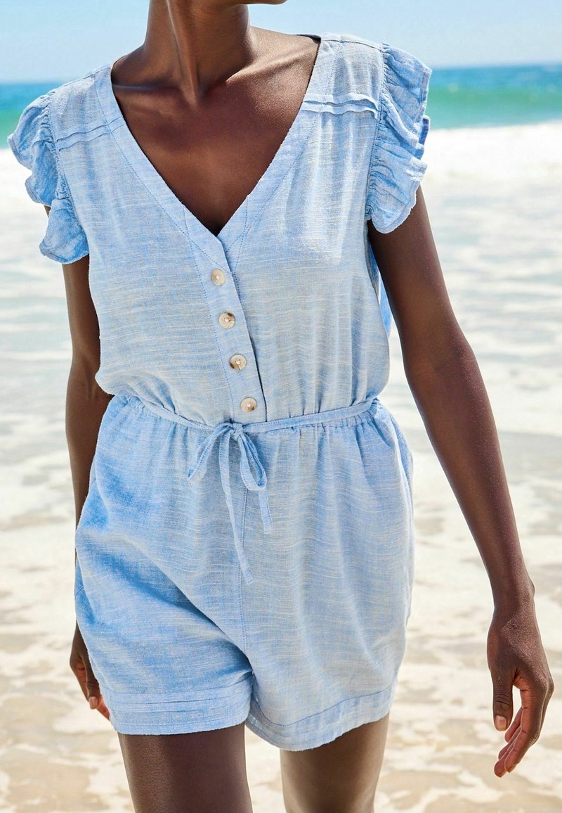 Femme portant une combinaison courte bleu clair à manches courtes avec des boutons et une ceinture nouée à la taille, debout sur une plage de sable près de l'océan.