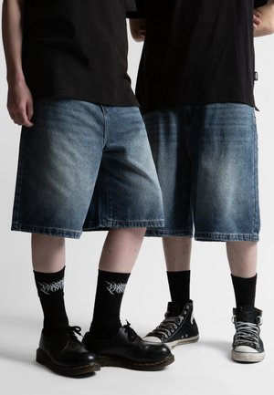 To personer står side om side iført sorte skjorter, lange denimshorts, sorte grafiske sokker og sort fodtøj.