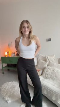 Ženska z dolgimi blond lasmi, oblečena v belo majico brez rokavov in črne hlače, stoji v prijetni dnevni sobi z zeleno omarico in belo zofe.