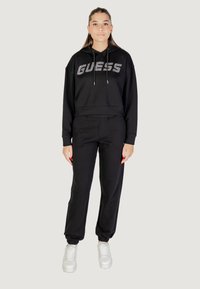 Sweat à capuche noir avec le logo "GUESS" en argent, associé à un pantalon de jogging noir et des baskets blanches ; coupe décontractée, tissu confortable.