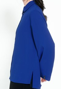 Blå, långärmad skjorta med krage, sidofickor och en slät yta. Vänster ärm har en taggdetalj. Enfärgad, inga mönster.