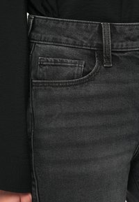 Tamsiai pilkos džinsinės kelnės, turinčios klasikinį penkių kišenėlių dizainą, matomus siūlus ir vidutinio aukščio liemenį su diržo kilpa.