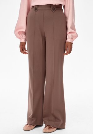 Hoge taille, brede poot bruine broek met knopen aan de voorkant, gedragen met lichtroze top met lange mouwen en bijpassende bruine ballerina's.