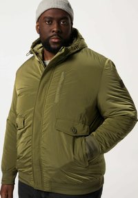 Giacca puffer verde oliva con zip frontale, due grandi tasche anteriori e polsini a coste. Presenta un cappuccio e un tessuto testurizzato.
