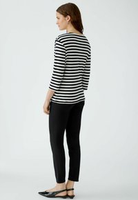 Schwarz-weiß gestreifter Langarm-Oberteil, Rückansicht, kombiniert mit schwarzen Slim-Fit-Hosen und schwarzen Slingback-Schuhen, vor einem minimalistischen Hintergrund.
